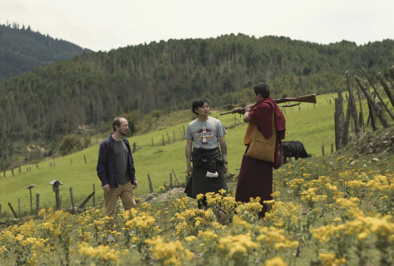 A white man and an Asian man stand in a field of yellow flowers, tree-covered hills behind them. They are talking to a Buddhist monk who has a rifle slung over his shoulder.