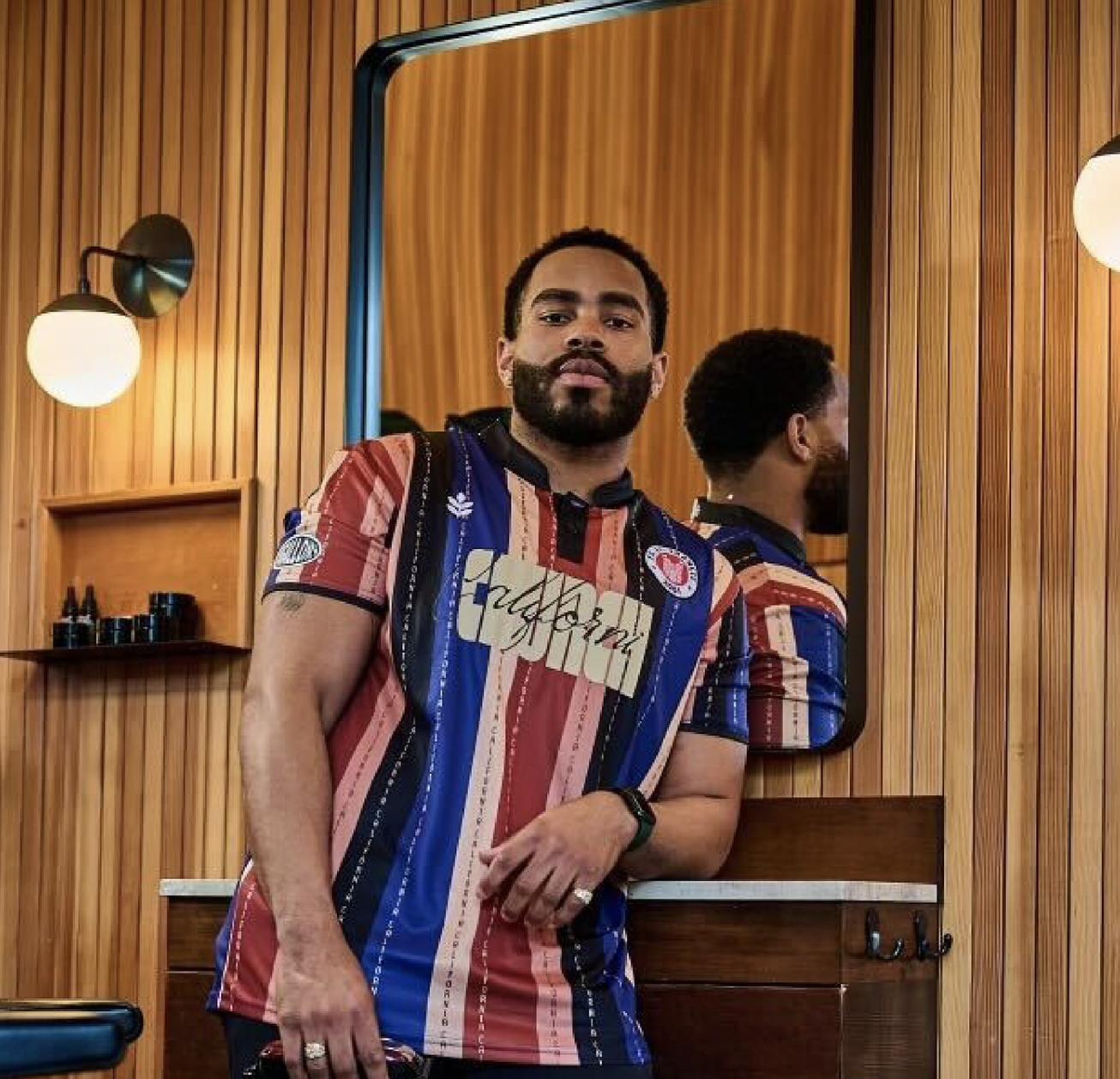 A Black man wearing a vertically striped shirt leans against a barber station with a mirror behind him.