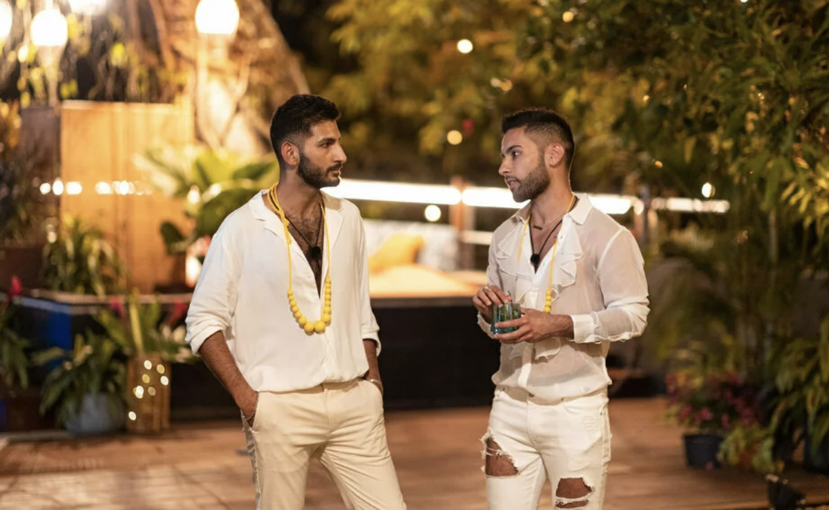 Two men stand in a tropical looking resort, both wearing white shirts and pants, both looking serious.