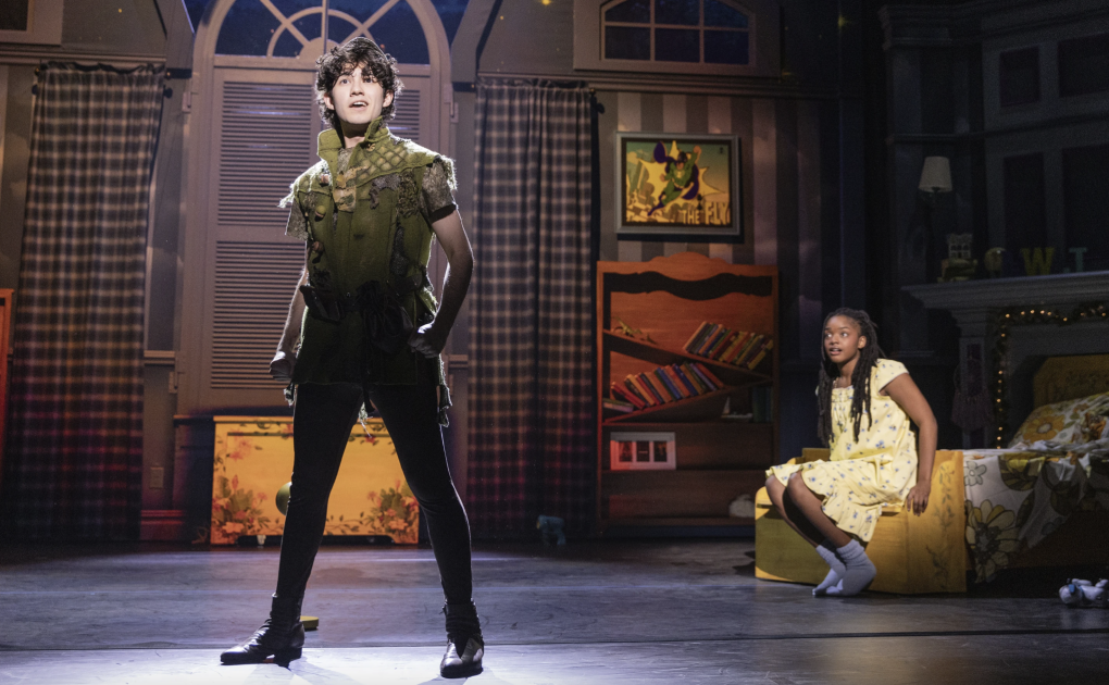 A stage set of a bedroom. Peter Pan stands front of stage, a Black girl sits behind, close to the floor.