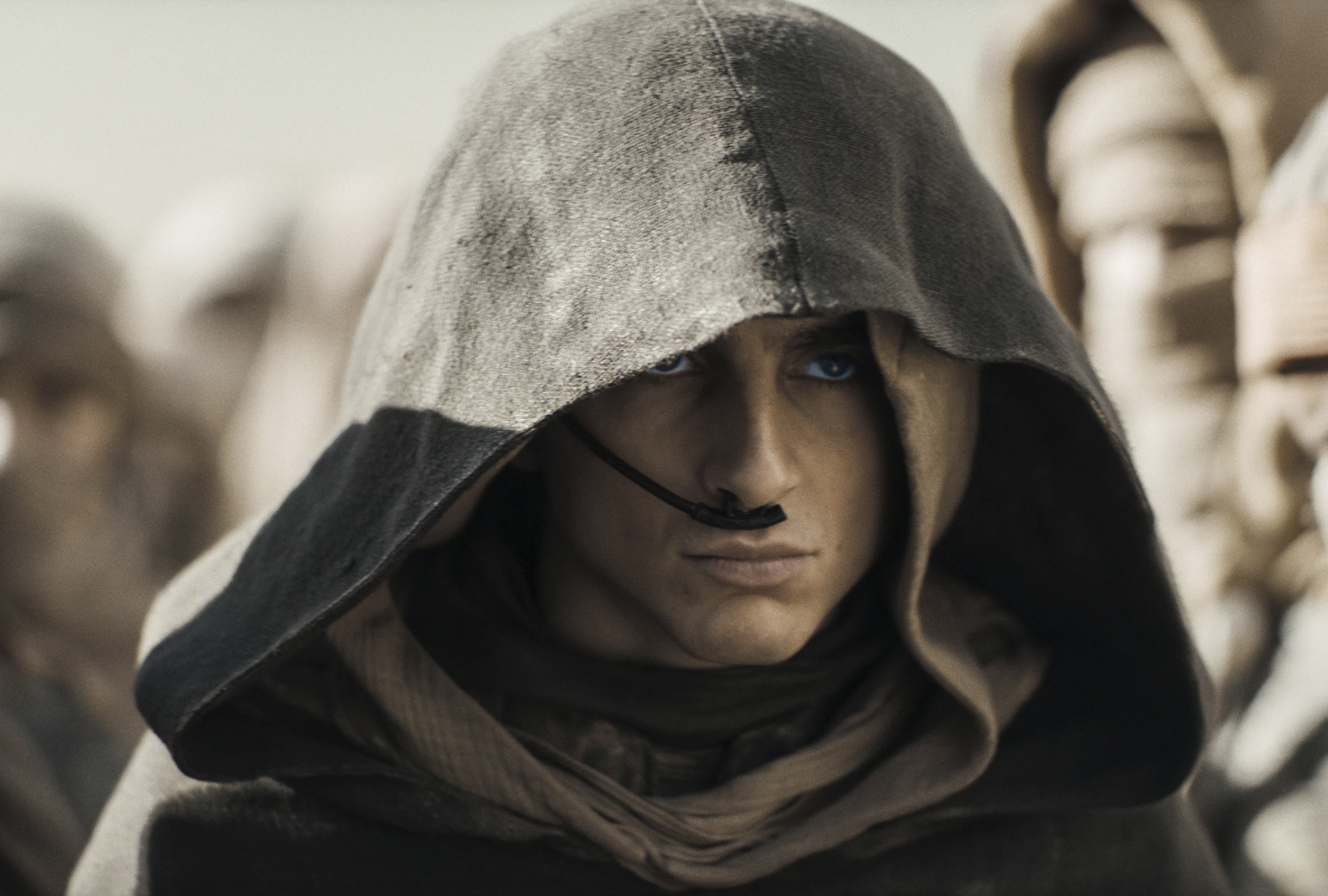 A mysterious white man peers out from under a dark hood. There is a small mechanism placed under his nose.