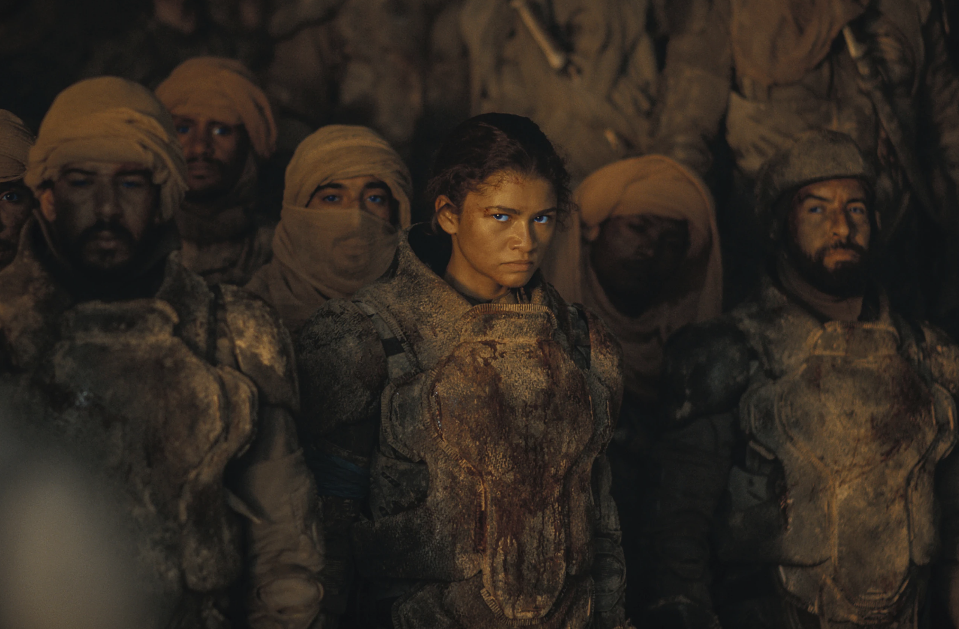 A woman glares into the distance with thinly veiled anger. She is wearing dirty utilitarian clothes and is surrounded by men dressed similarly.