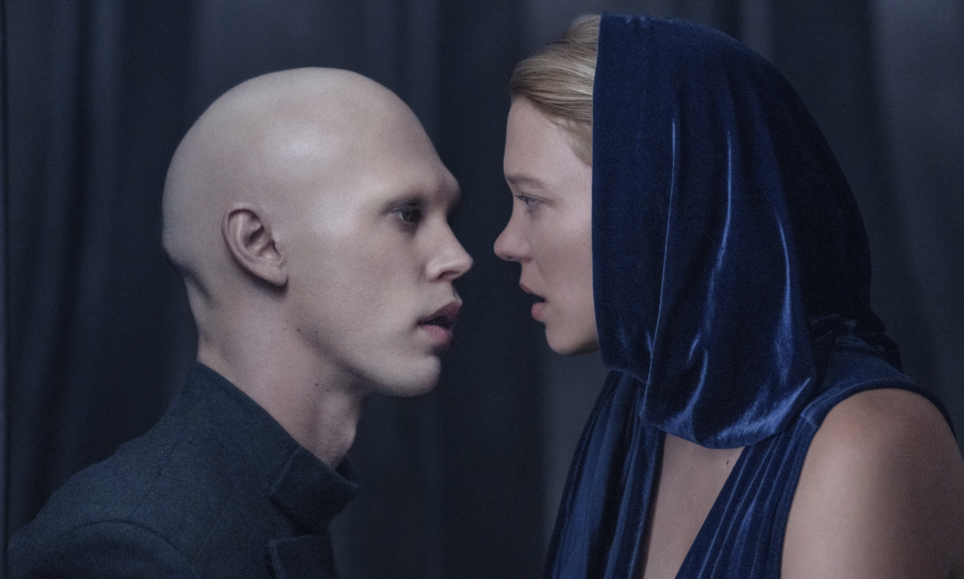 A pale young white man with a bald head and no eyebrows gazes at an attractive blonde woman wearing a blue hooded velvet dress. Their faces are close together.