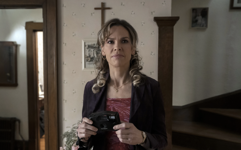 A middle-aged white woman stands in a home and holds a camera. On the wall behind her is a cross.