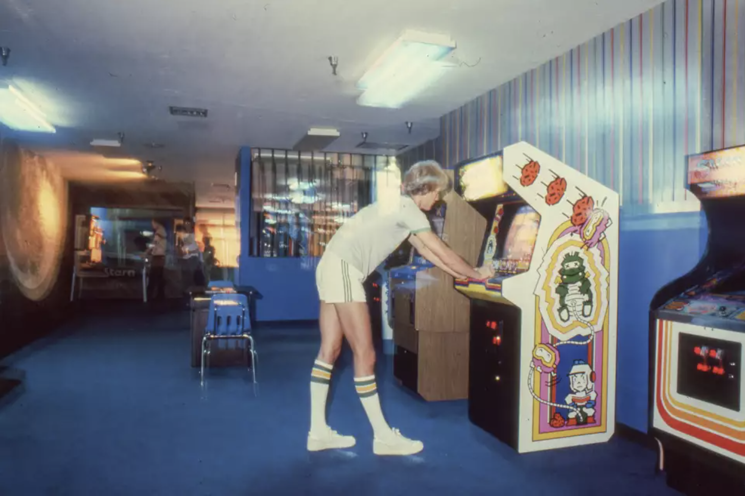 A young white man wearing short shorts and knee high tube socks plays an arcade game.