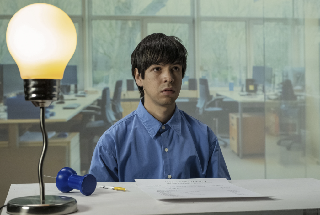 A youthful Latino man wearing a blue shirt sits at a table next to a lightbulb-shaped lamp.