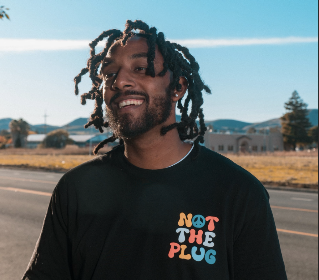 LOE Gino, posing for a photo while wearing a black shirt that reads "NOT THE PLUG" in colorful writing over his left chest area.