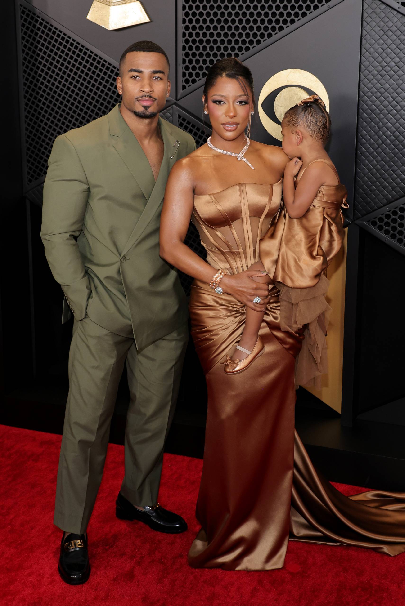 A Black man in a green suit and a Black woman in a tight rose gold gown stand on the red carpet. The woman is holding a small child.