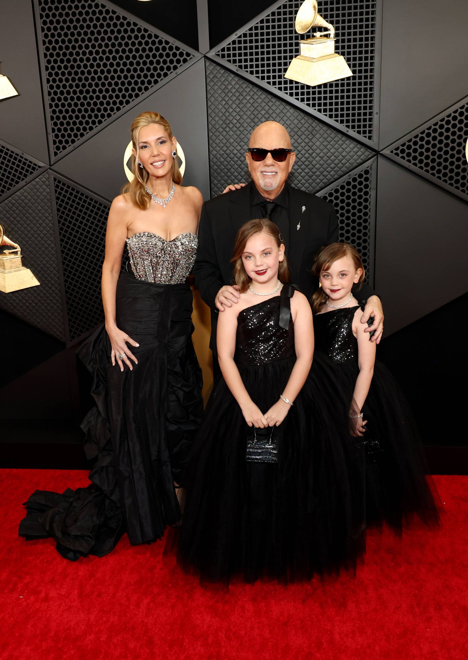A slender blond woman in a black and silver dress and a bald man in black suit and sunglasses stand on the red carpet with two young girls in black dresses.