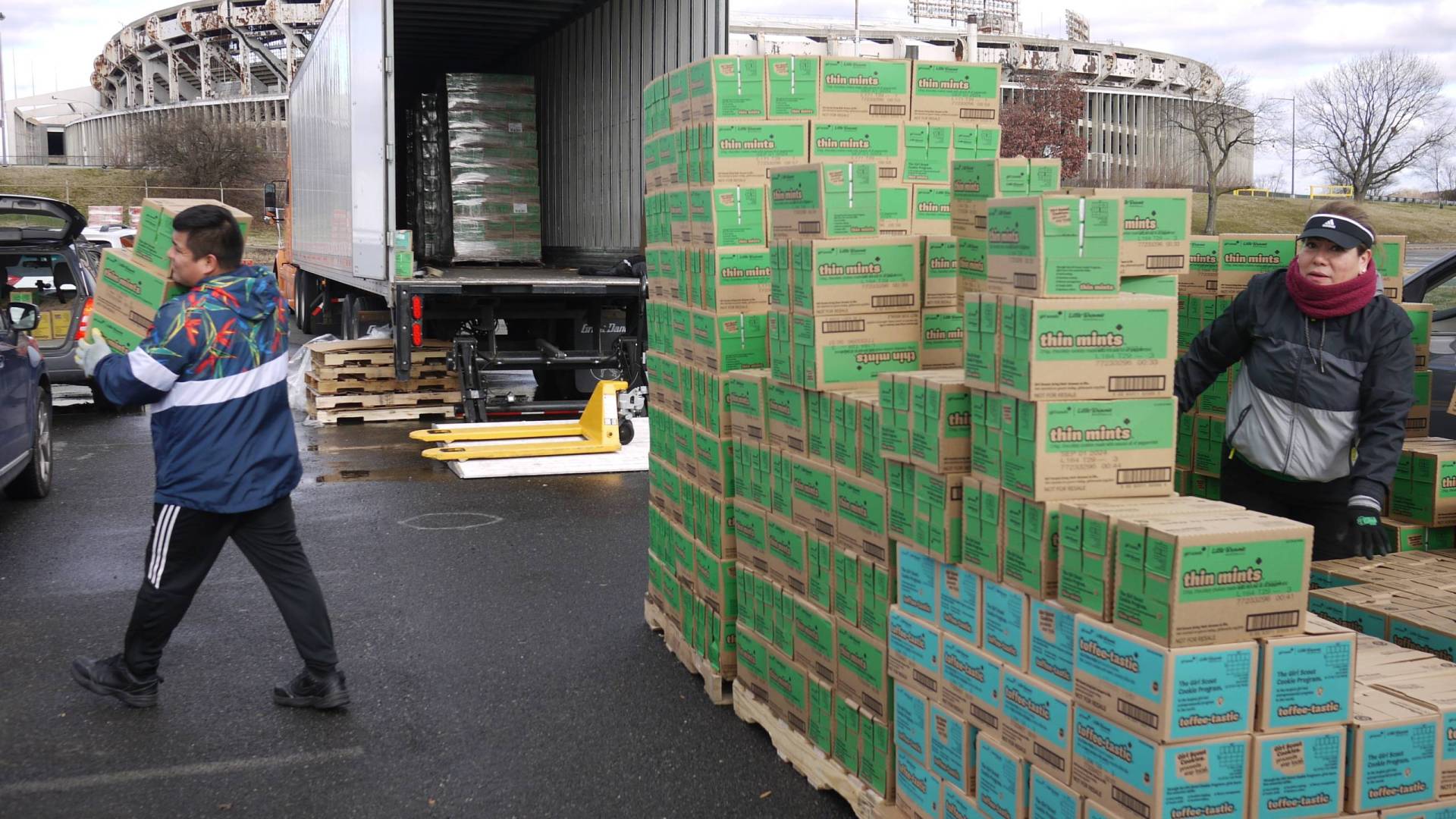 A man carries a pile of green and brown boxes to a vehicle, while a woman dressed for winter stands behind a wall of the same boxes.