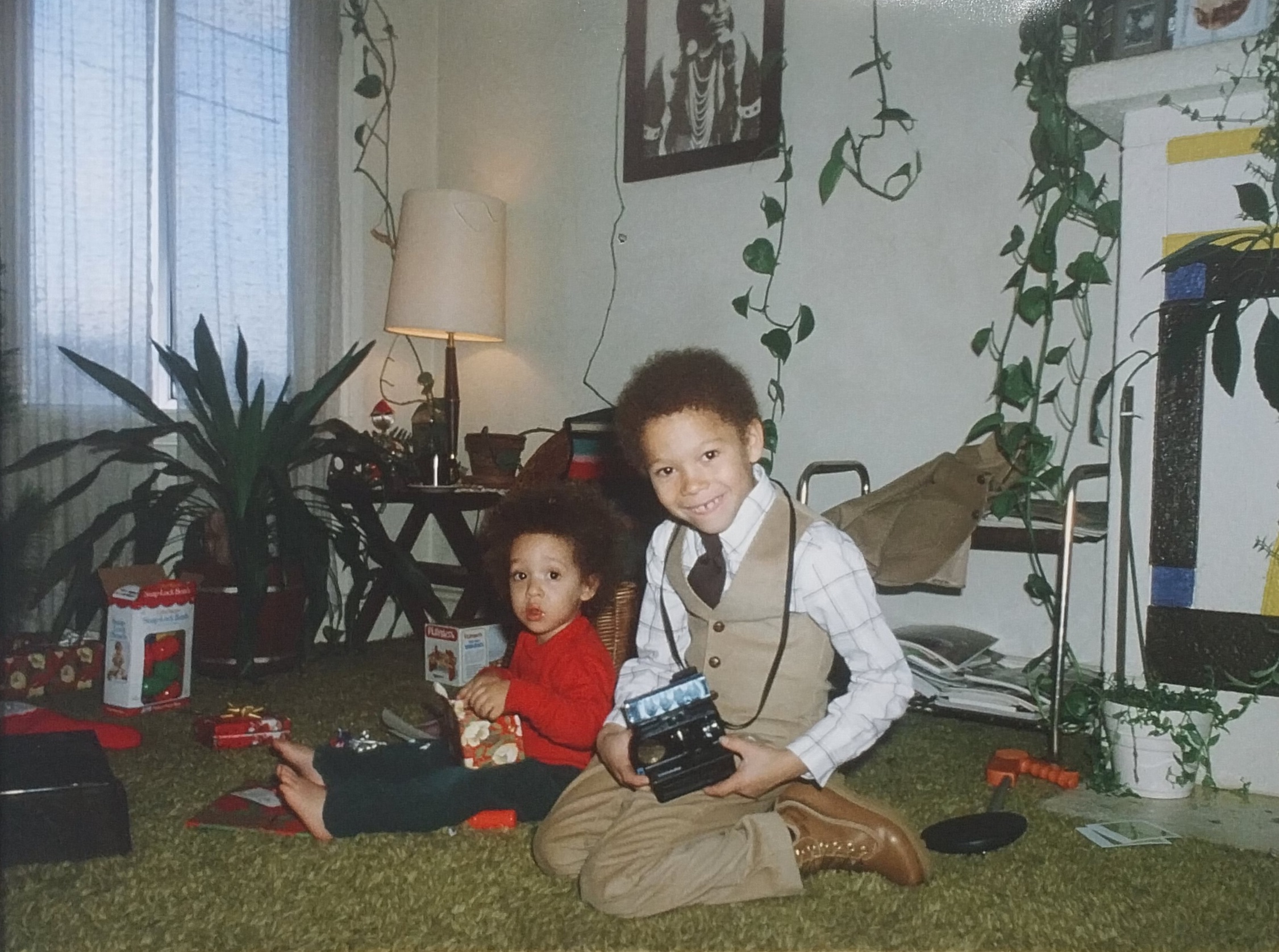 Two children sit on the floor of a comfortable living room. They are surrounded by green plants, a white lamp behind them. 