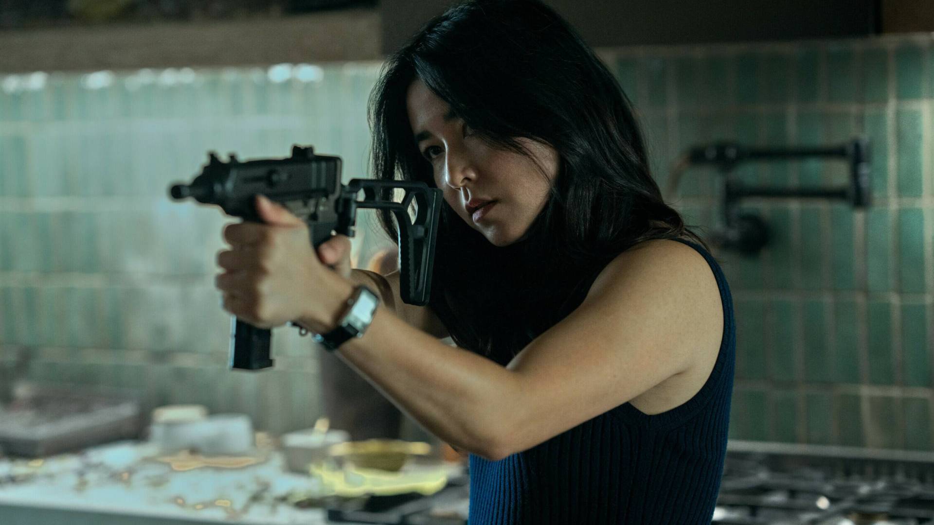 A Asian woman stands n a kitchen holding up a large automatic weapon.