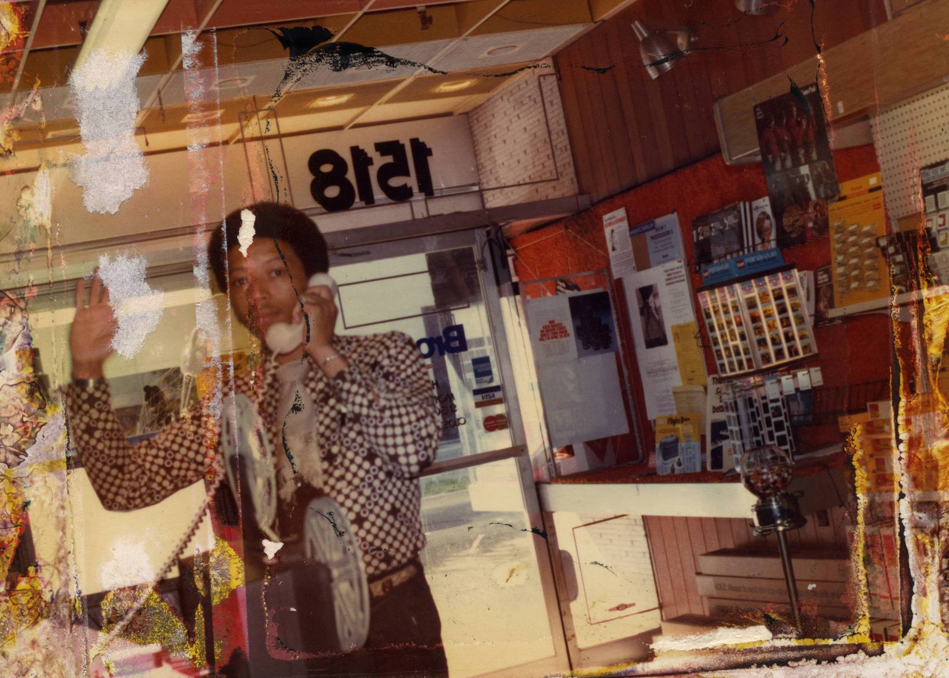A slightly damaged photograph of a Black man on the phone inside a small business. He is waving to the photographer.