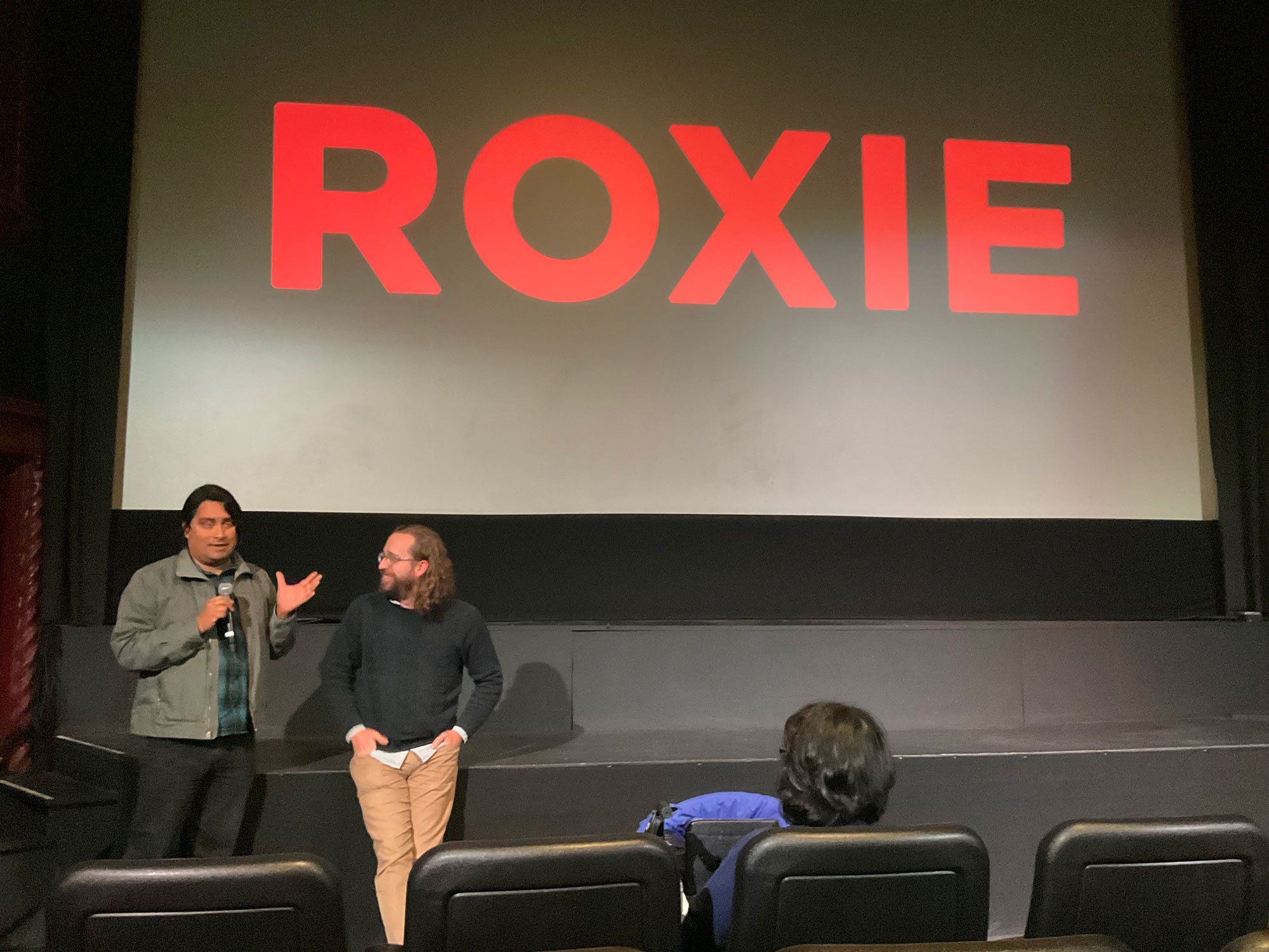 Two people lean against stage facing theater seats with big 'ROXIE' in red on screen