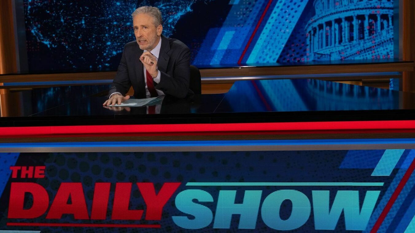 A white man in a suit sits behind a news desk in a TV studio.