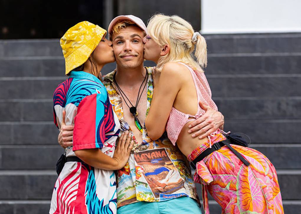 A man in brightly colored clothes stands with his arms around two women. Both women are kissing him on the cheeks.