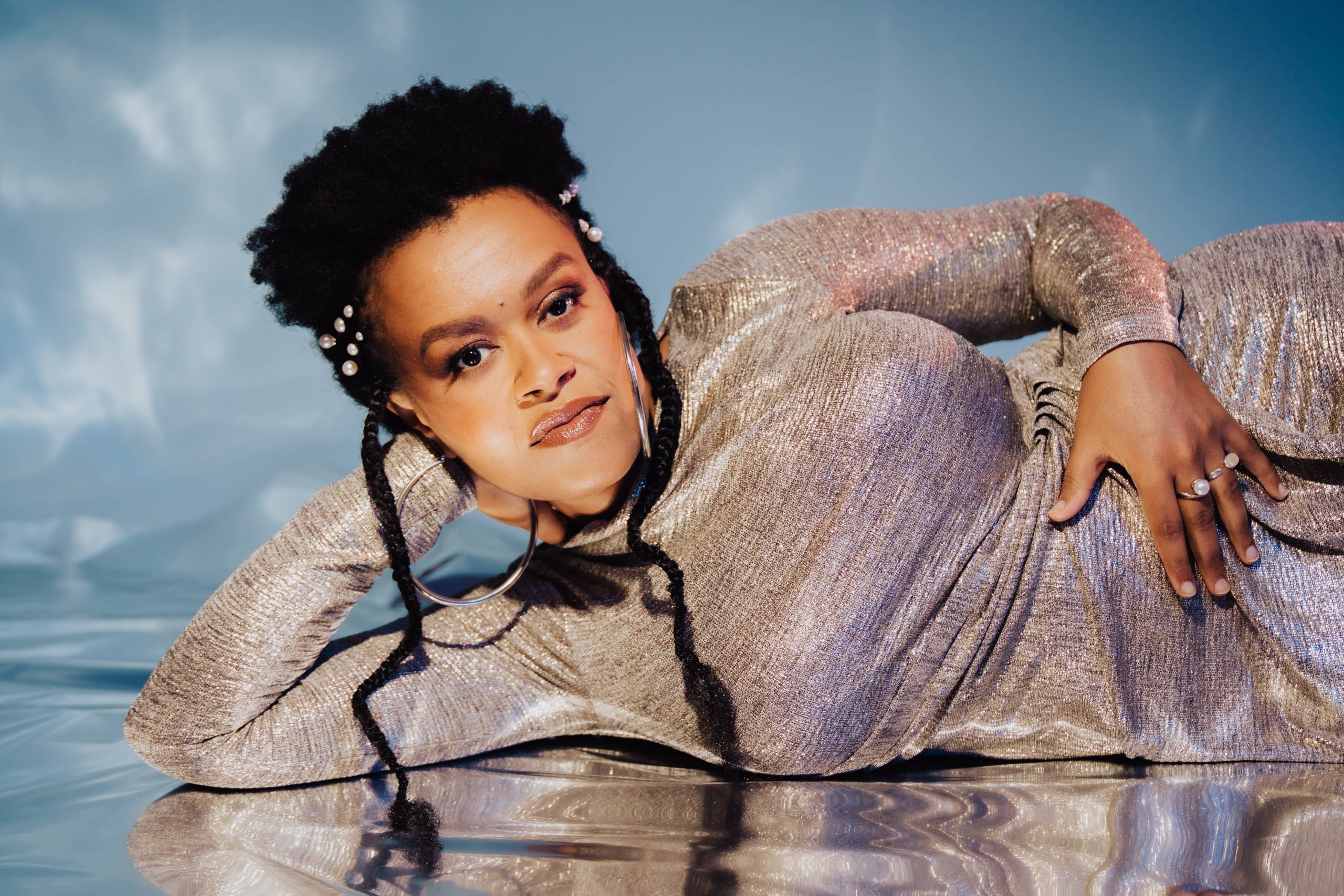 a Black woman in a silvery dress poses lying on a mirrored floor, propped up against one arm, looking at the camera