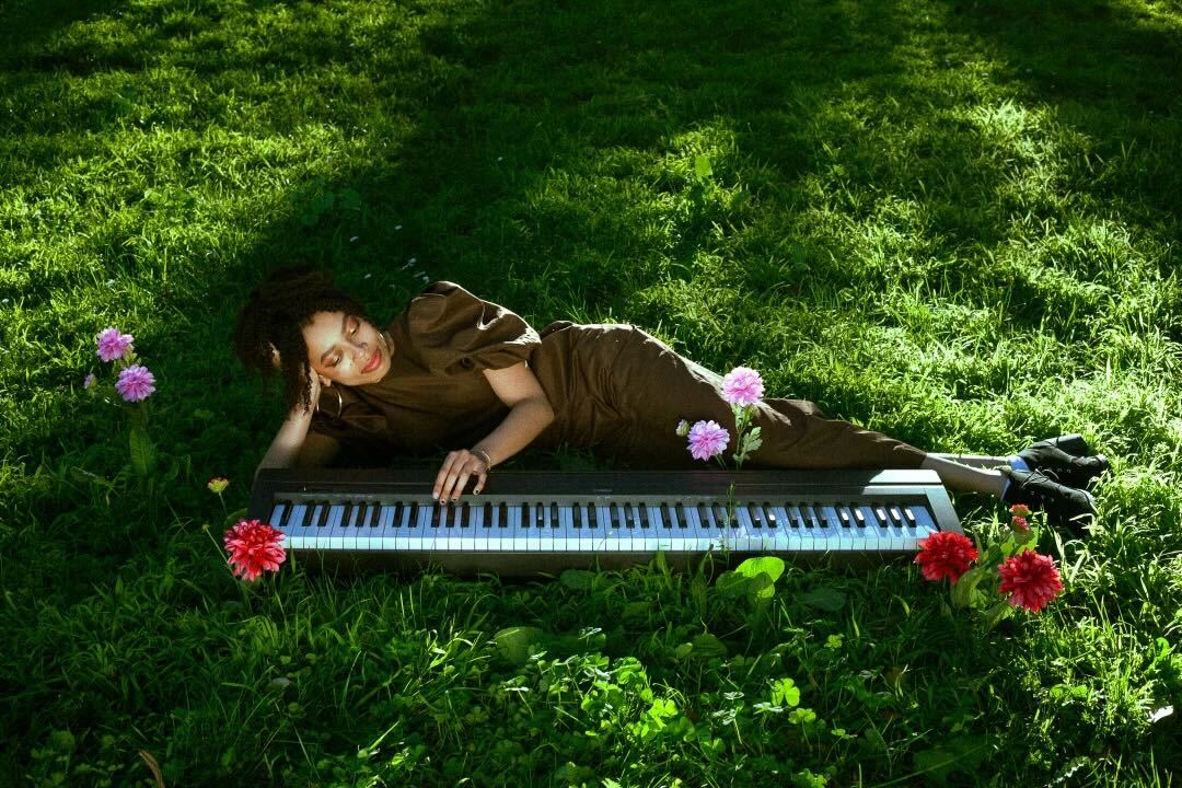 Singer and musician August Lee Stevens lays in a field of grass next to her full-sized keyboard.