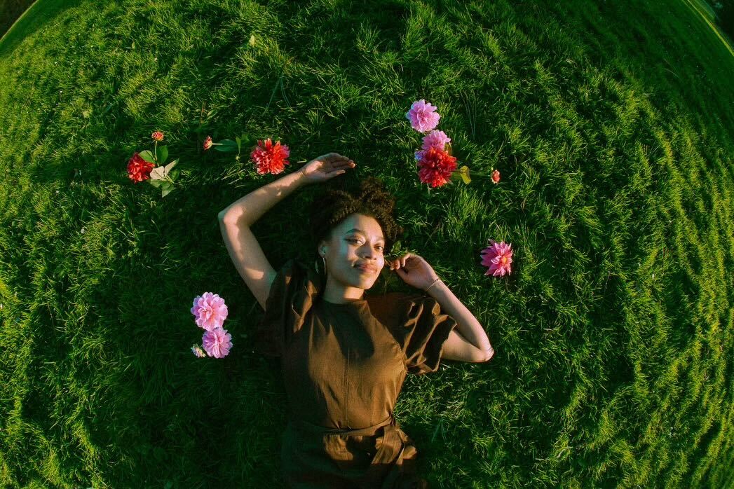 A young Black woman pictured from above, laying in a field of grass and surrounded by flowers, photographed with a wide-angle lens.