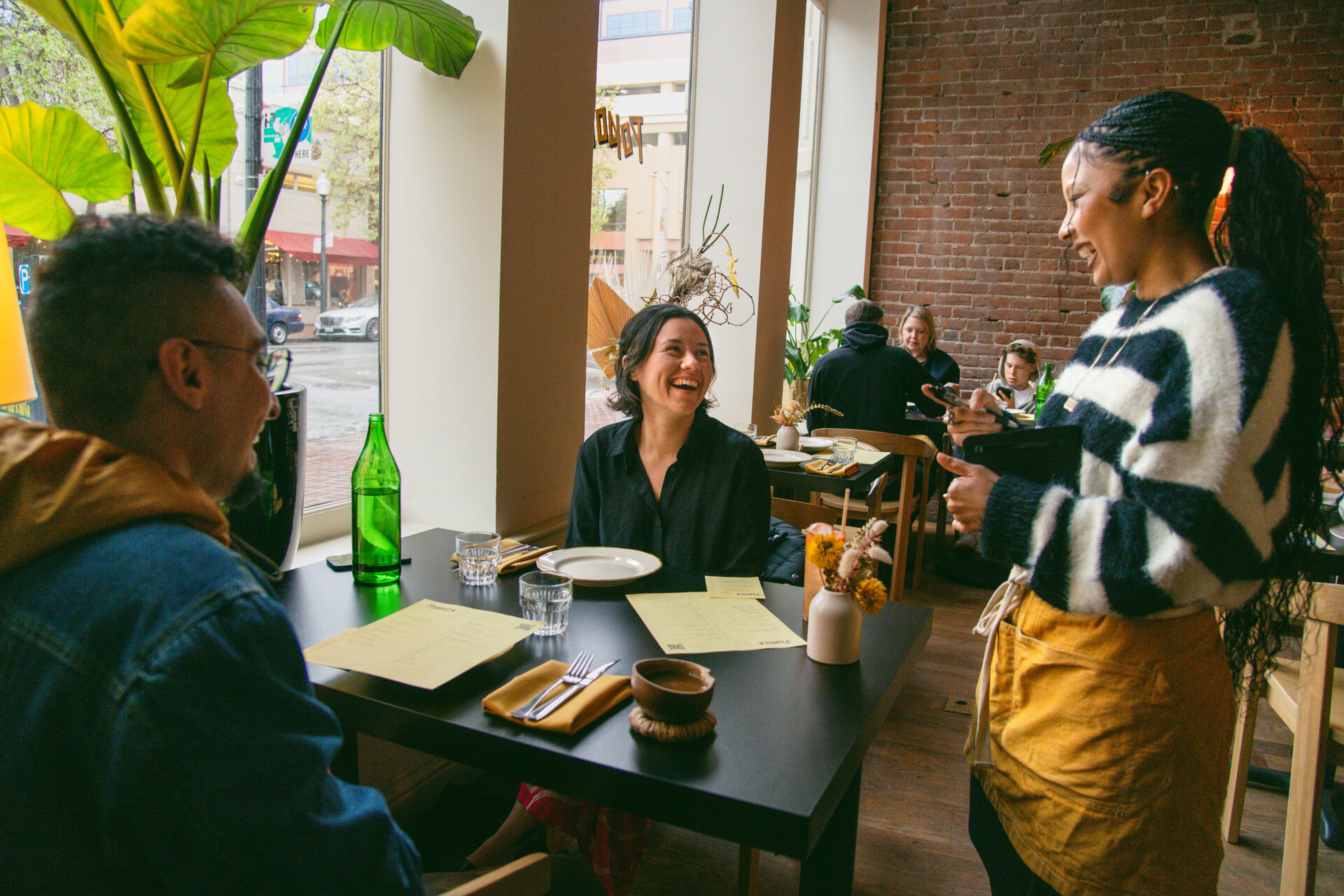 A restaurant server laughs as she takes an order from two customers sitting inside a sunny restaurant dining room.