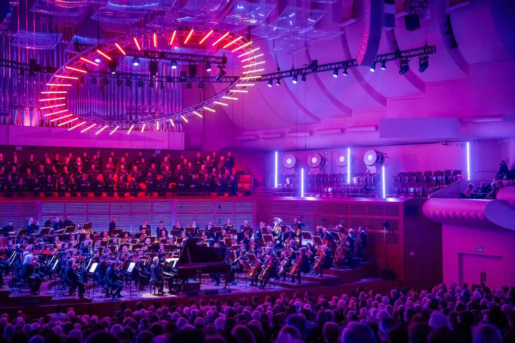 A circle of red lights hovers above a symphony orchestra, bathed in indigo hues, inside a concert hall