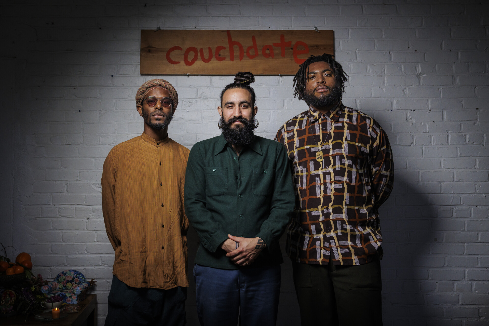 Three men stand facing the camera in front of a wooden sign that read "couchdate"
