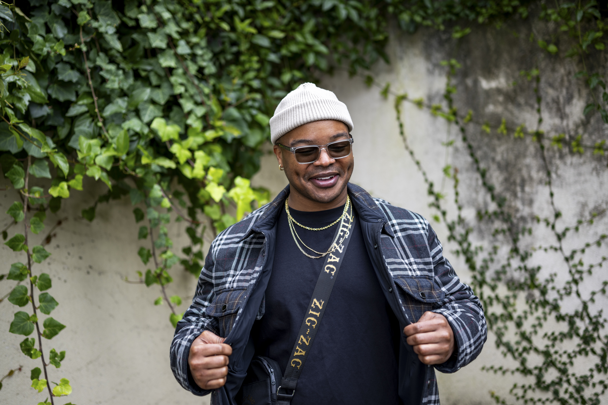Person in white beanie, plaid jacket, dark shirt, cross-body bag opens jacket and smiles