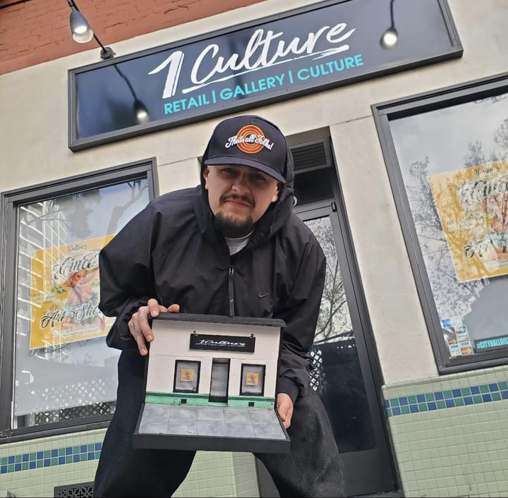 A man in a black hoodie and cap holds a small diorama of an art gallery storefront, standing in front of the same art gallery in real life