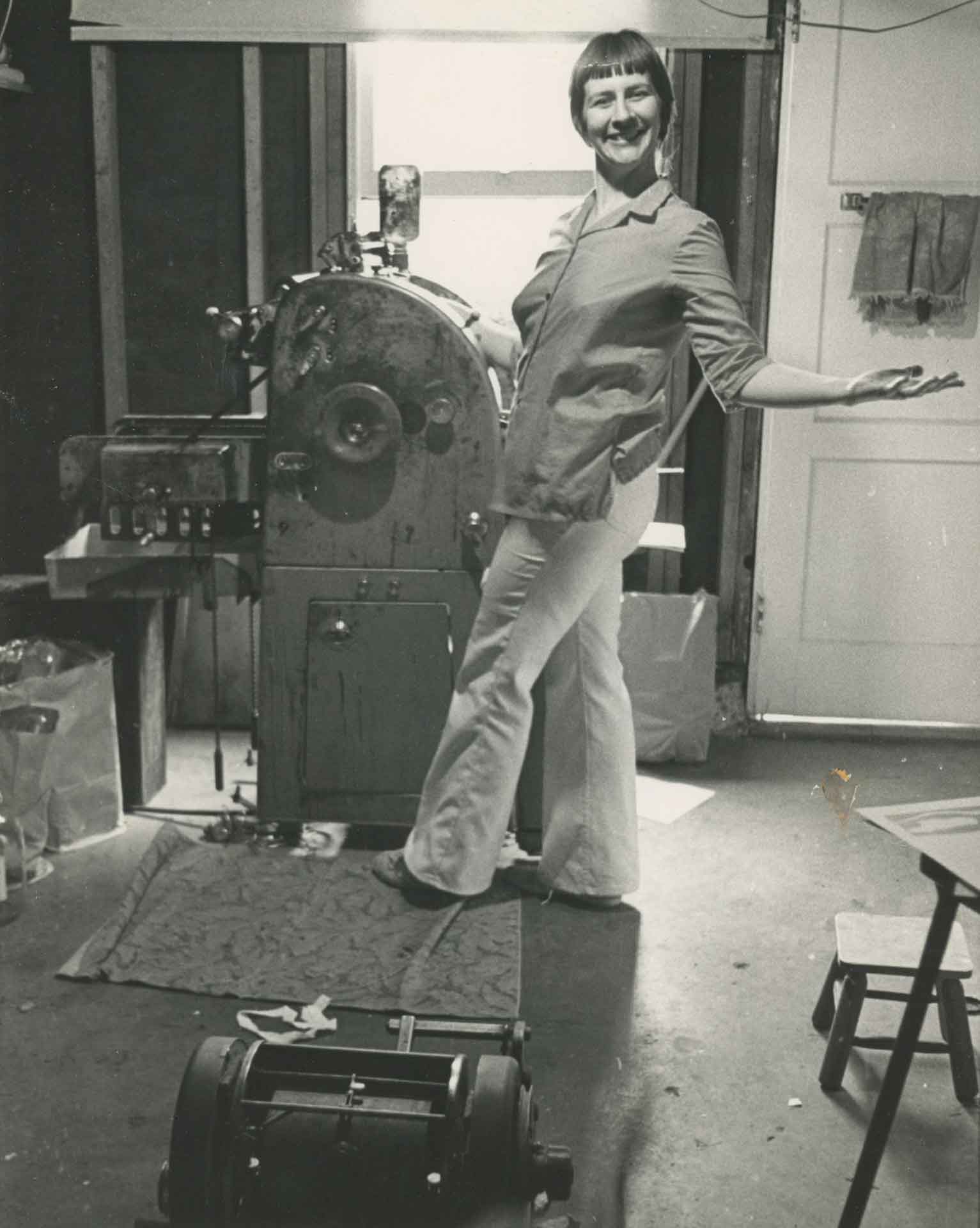 A young woman with a short bob haircut stands in a collared shirt and pants at a large metal printing press in a cluttered room.