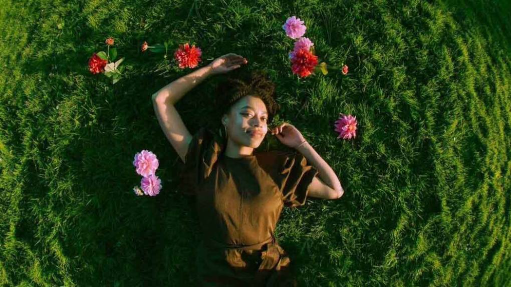 A young Black woman pictured from above, laying in a field of grass and surrounded by flowers, photographed with a wide-angle lens.