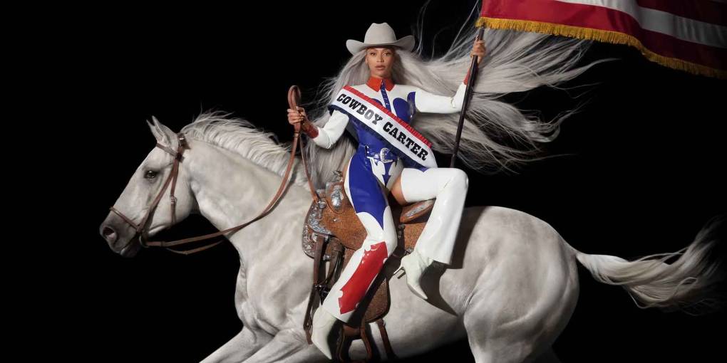 A woman in red white and blue clothes, with a sash reading 'Cowboy Carter,' sitting side saddle on a horse and holding an American flag