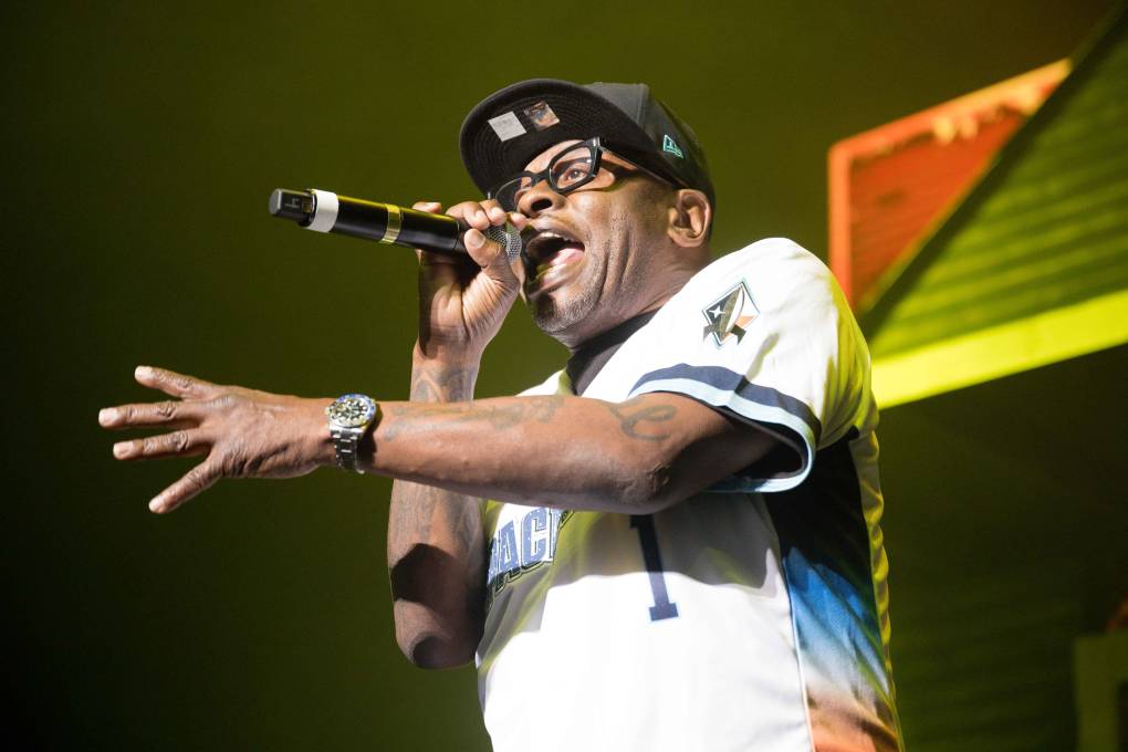 A Black man in baseball jersey and cap is seen from below, rapping into a microphone