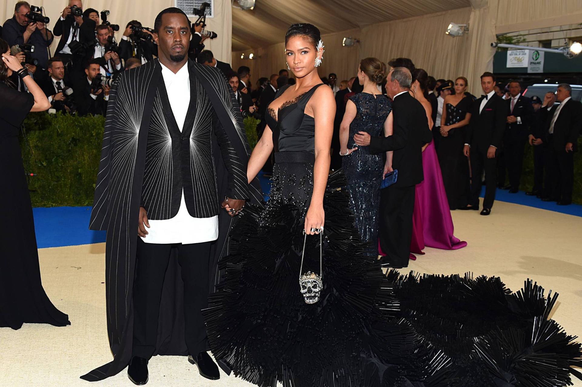 A Black man wearing a suit and very long couture coat stands holding hands with a Black woman wearing an elaborate black gown and purse in the shape of a skull.