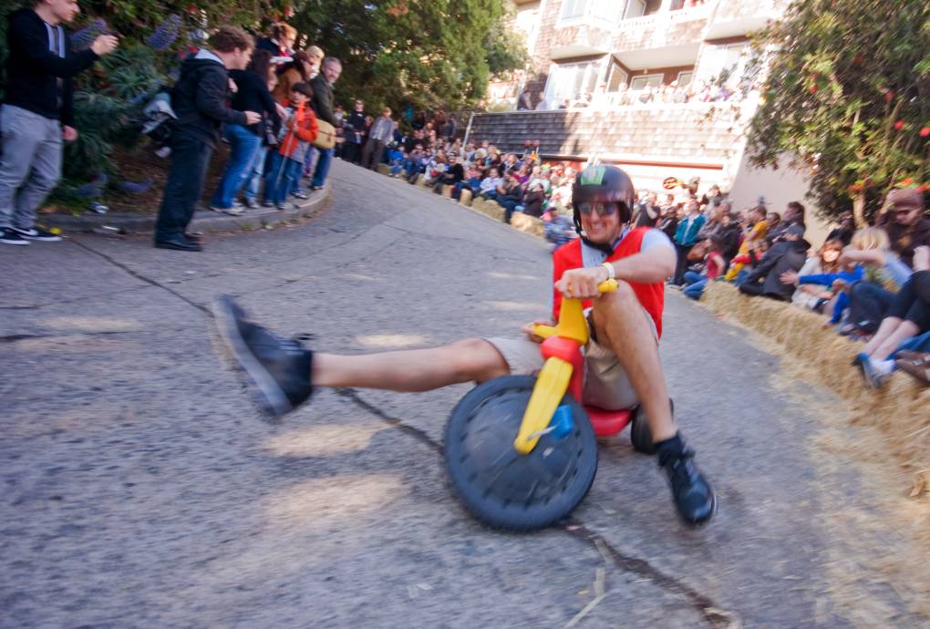 An adult riding a children's tricycle extends one leg as he takes a steep turn. He is wearing a helmet.