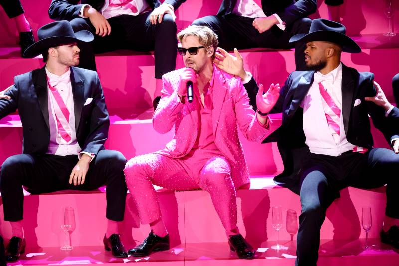 A man in a pink suit and shirt sits on pink stairs and sings into a microphone, surrounded by a group of men in black suits.