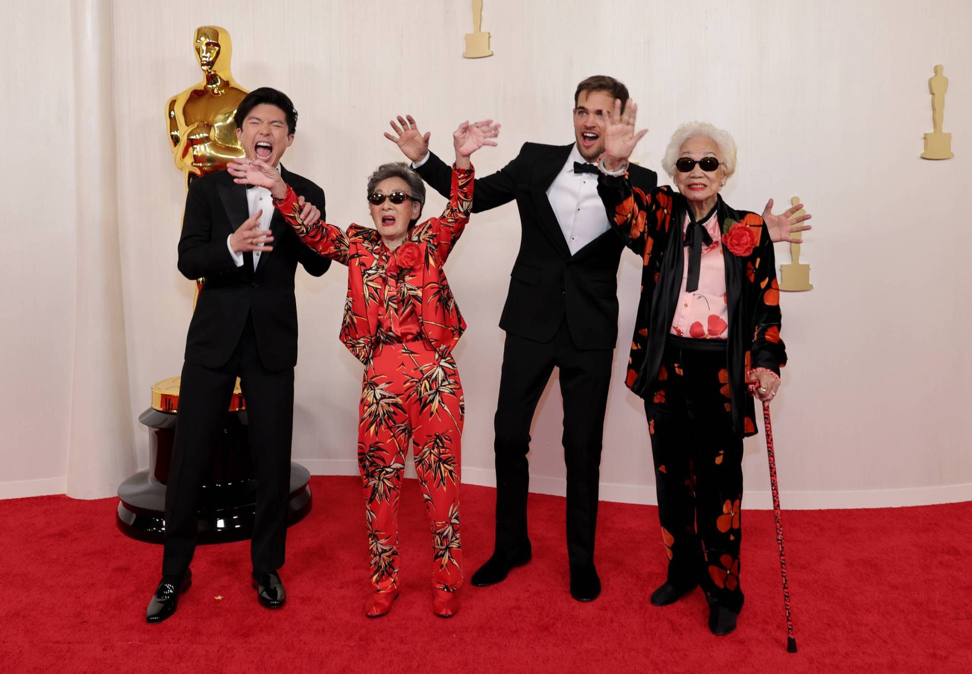 A white man and an Asian man, both wearing tuxedos gesture wildly next to two elderly Asian women, both wearing dark sunglasses and flamboyant suits.