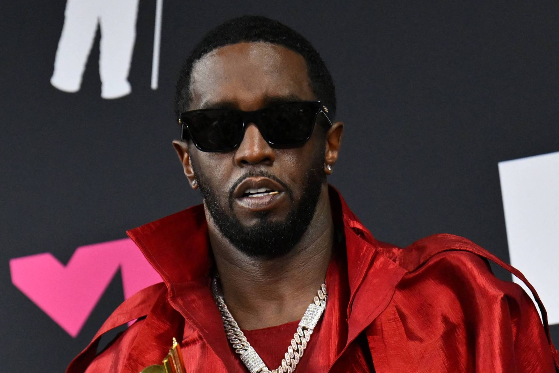 A Black man wearing dark sunglasses and a red coat faces the camera, visible from the shoulders up.