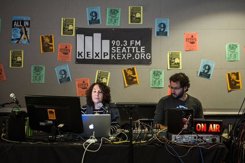 A radio host and engineer sit in a studio in front of a sign that reads "KEXP Seattle."