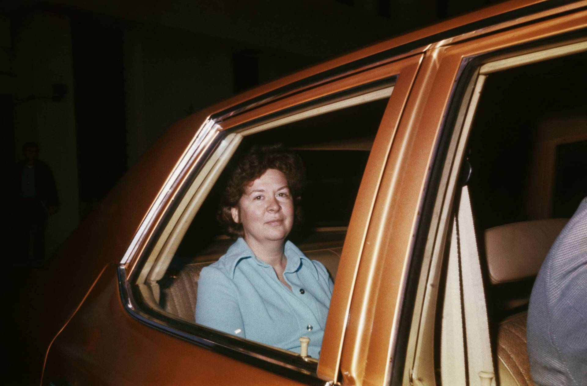A white woman in pale blue 1970s-era clothing sits in the rear of a brown car.