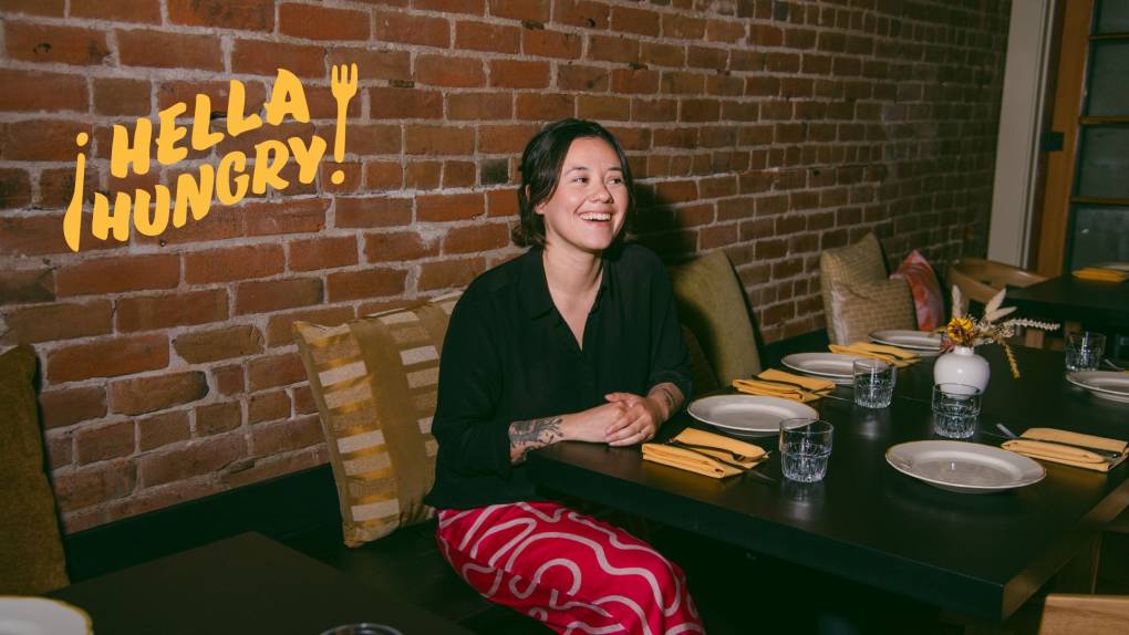A woman smiles sitting a a long wooden table inside a stylish restaurant with a red brick interior.