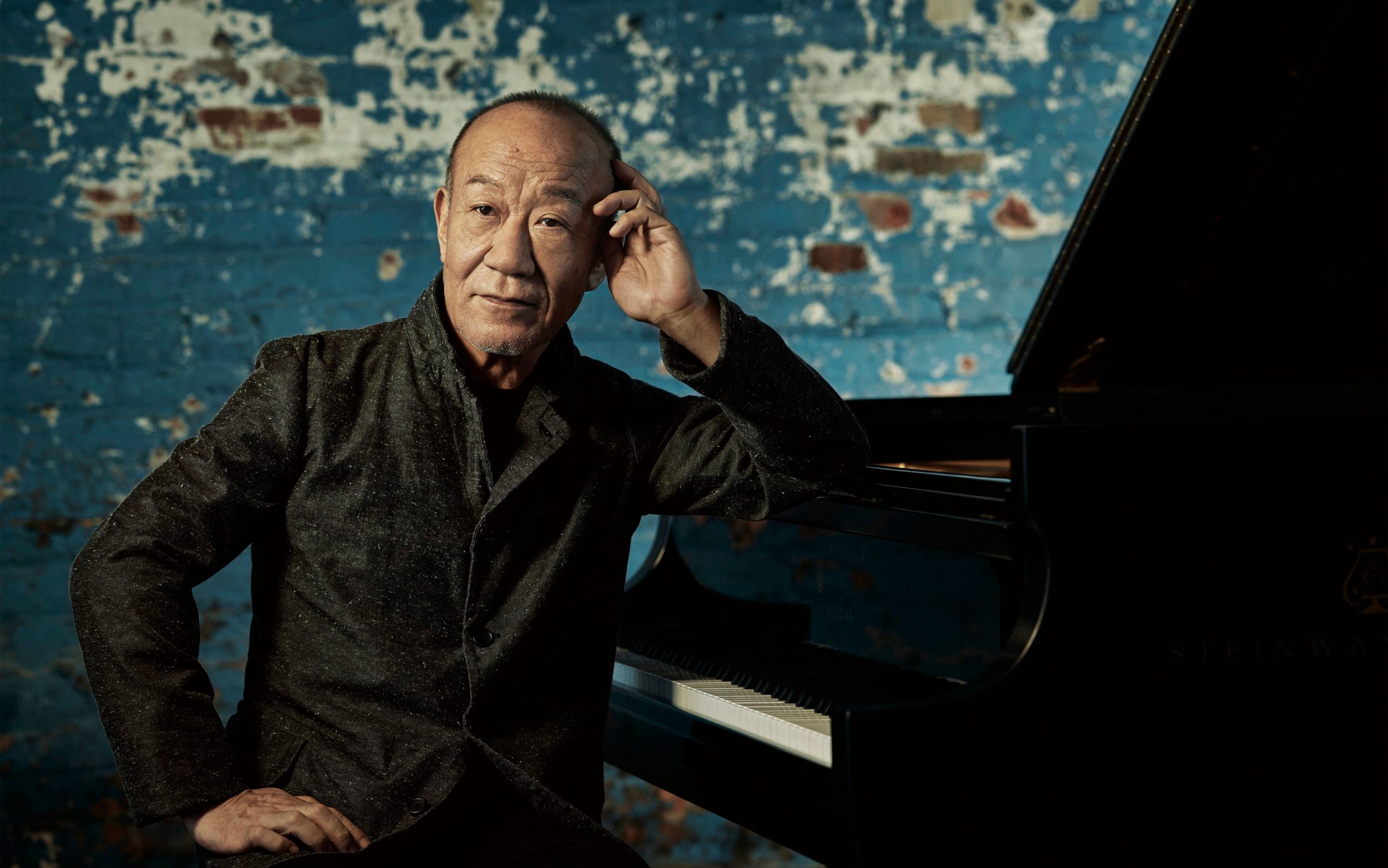 A Japanese man sits at a piano with an abstract pattern on the wall in the background