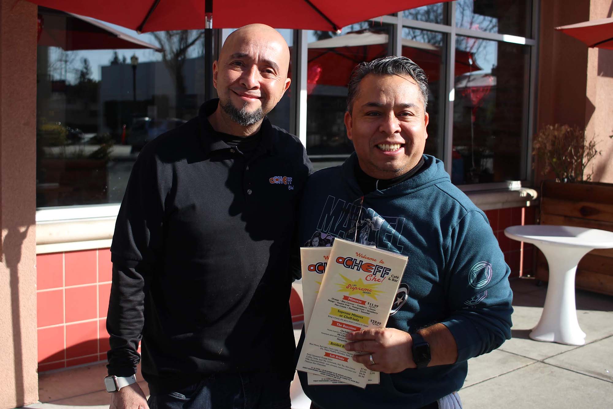 Two chefs pose outside of their restaurant; the one on the right holds a stack of menus.