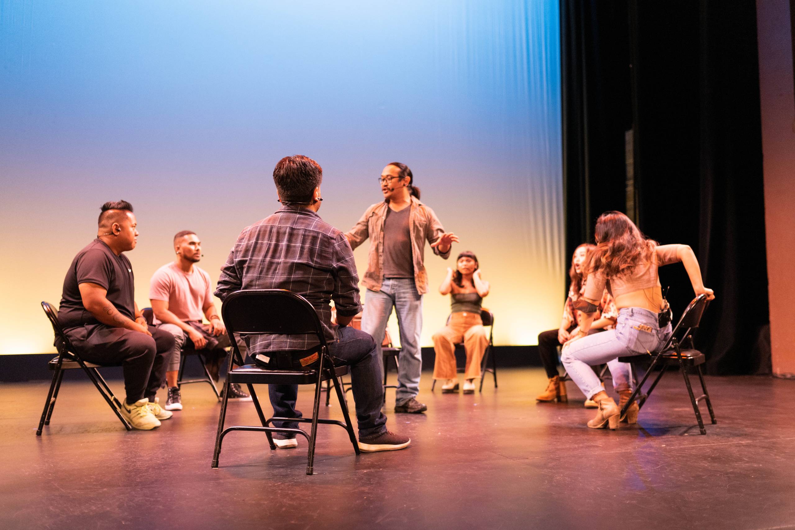 A man stands in the middle of circle of people seated in folding charges on a stage.