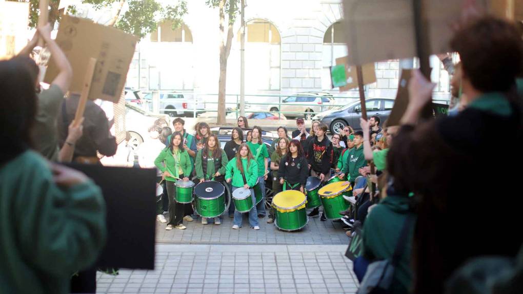 A crowd of drummers in green on the sidewalk is seen through the silhouetted backs of protesters holding signs