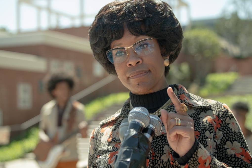 A still from the film 'Shirley,' where Regina King is in character as Shirley Chisholm, speaking into a microphone at a press conference.