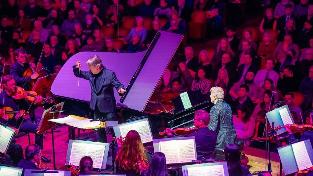 A man conducts an orchestra aside a pianist at a grand piano, on stage, bathed in purple light
