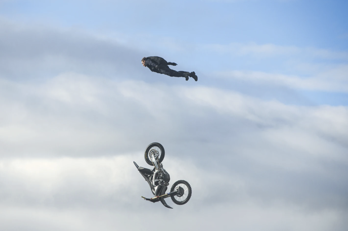 A man flies through the air in a horizontal position, a motorbike if falling vertically underneath him.