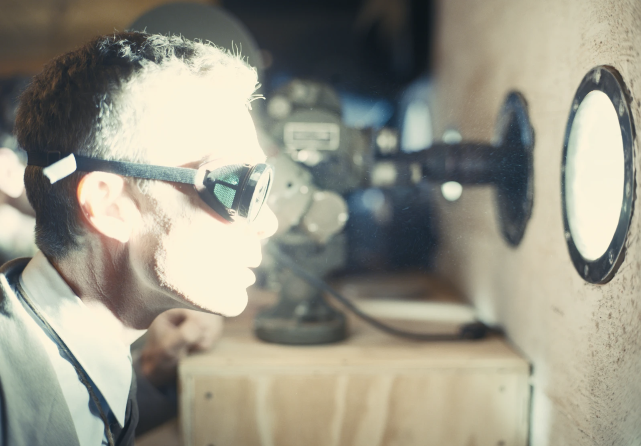 A white man sits, wearing goggles in front of a small window. His face is lit up in bright light.