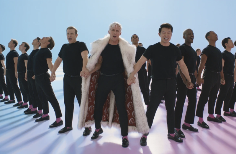 A white man in a long fur coat and an Asian man stand in a long line of men in matching black outfits, all holding hands in a line.