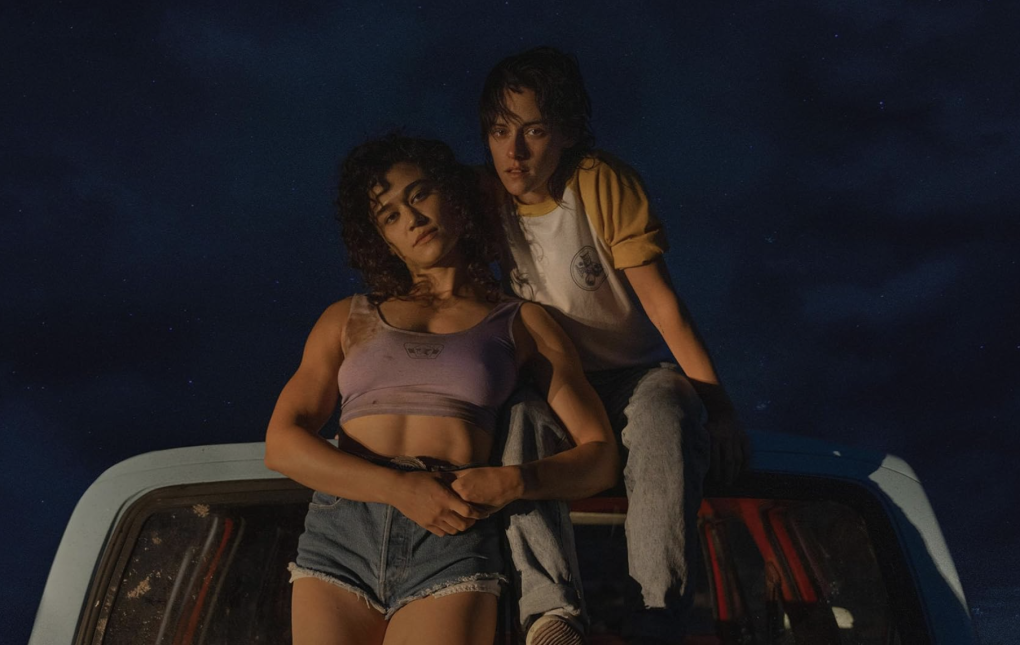 A muscular woman in cropped top and short shorts leads against a tomboyish woman sitting on top of a truck.